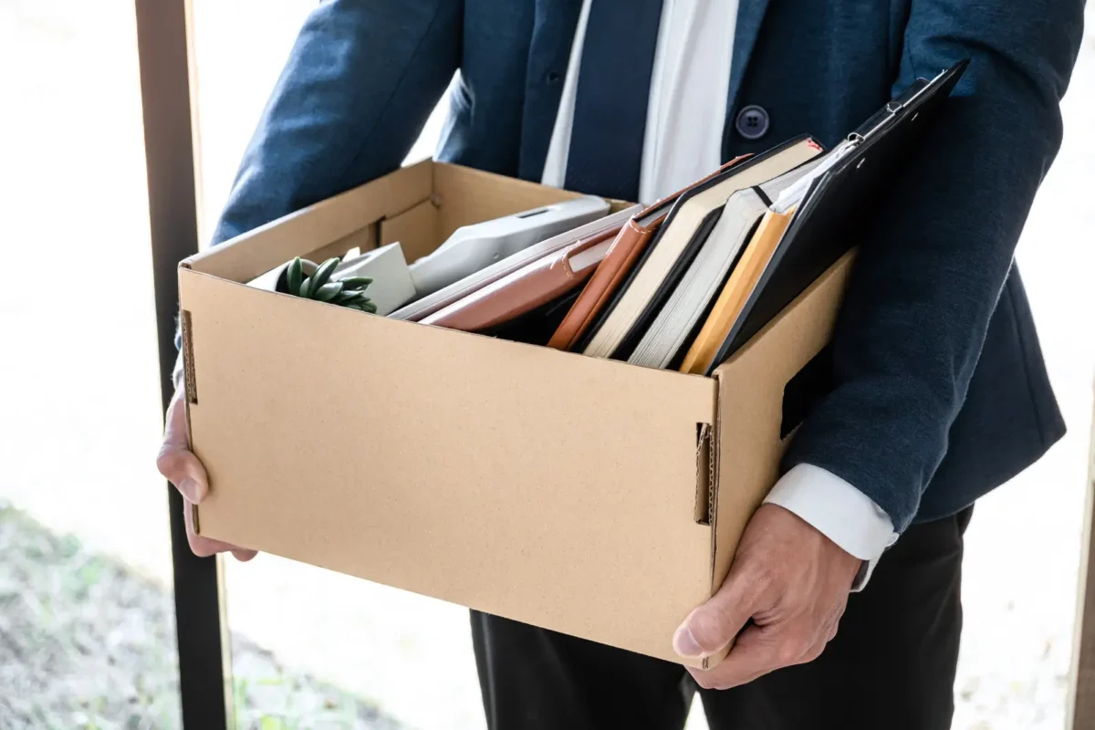 Employé en costume quittant son entreprise avec un carton contenant ses affaires après un licenciement.